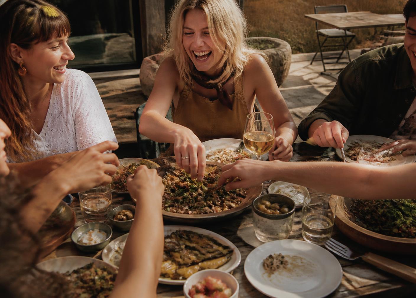 Friends laughing and sharing plates of food at a sunlit outdoor table